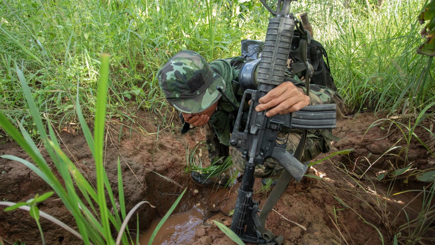 A soldier holding a gun crouching in the grass with his face obscured by his helmet